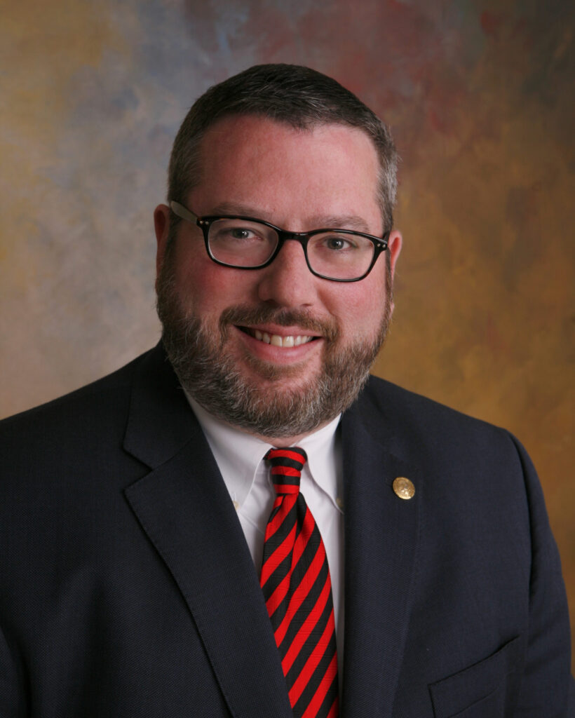 A man with short brown hair and glasses wearing a blue blazer, white collared shirt and navy and red striped tie