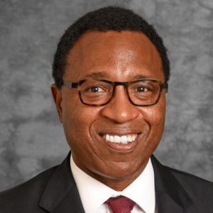 A man with short black hair and glasses wears a black blazer, white shirt and maroon tie
