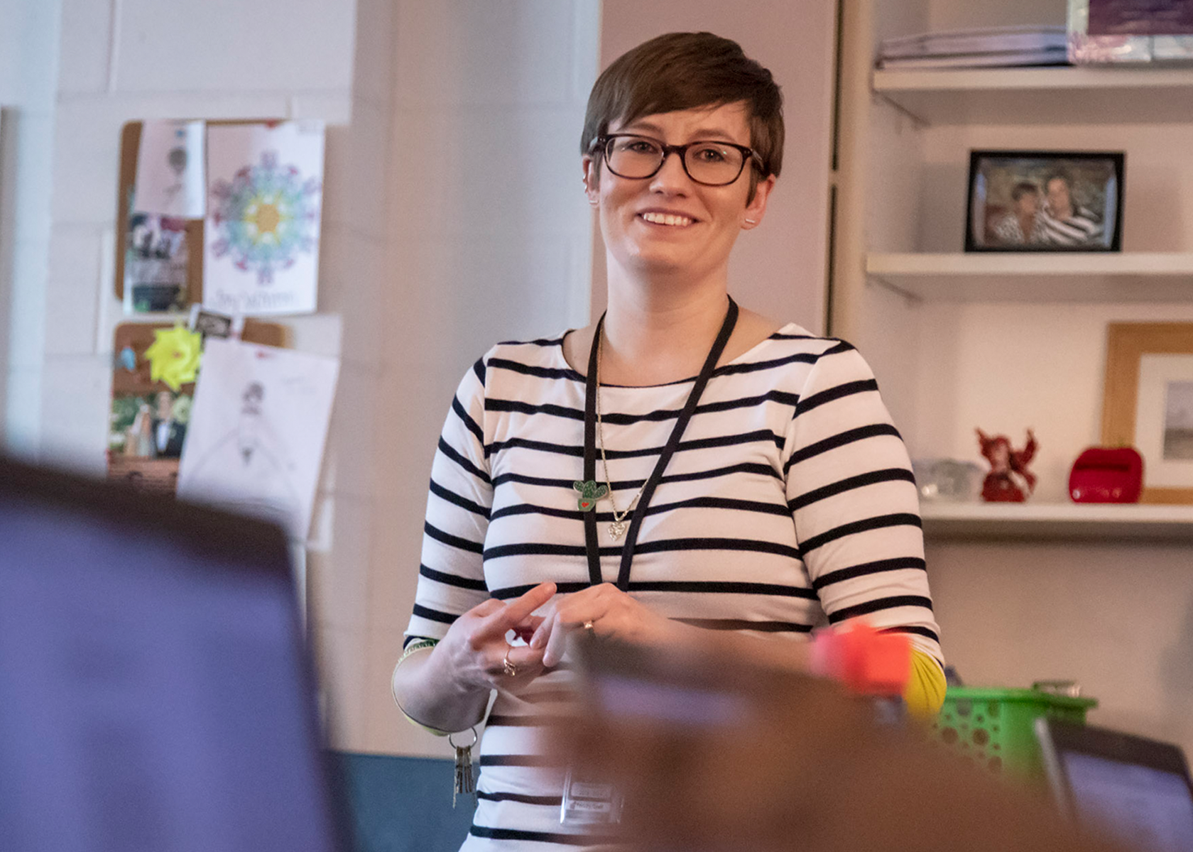A teacher stands in front of a classroom smiling. She has short brown hair and is wearing a lanyard over her striped shirt