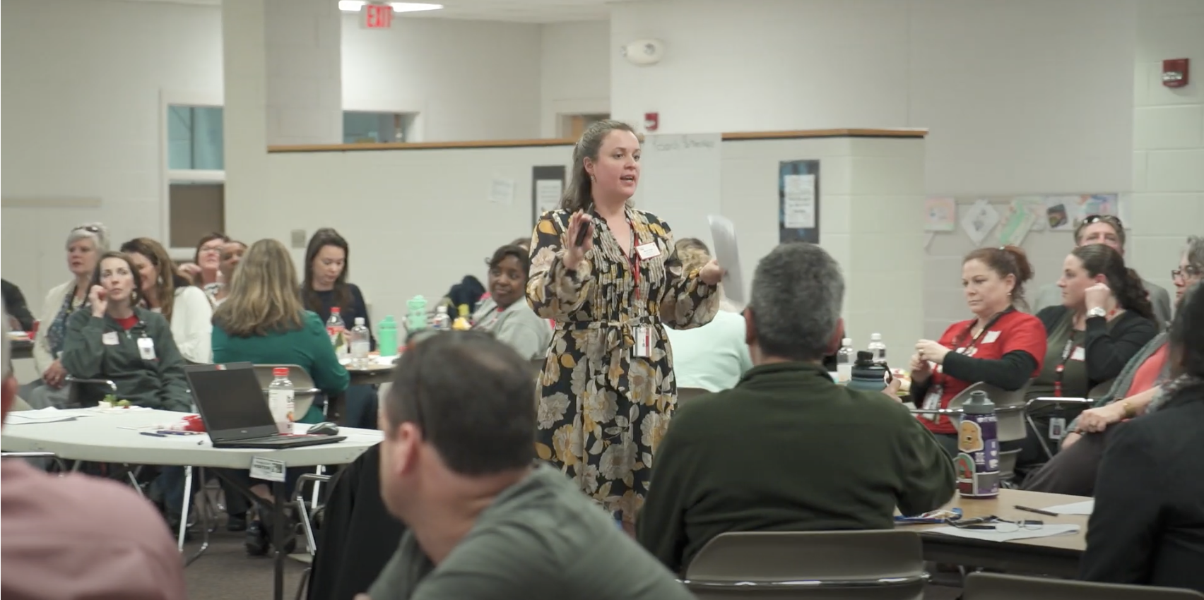 A woman speaks to a group of teachers sitting at tables situated around the room
