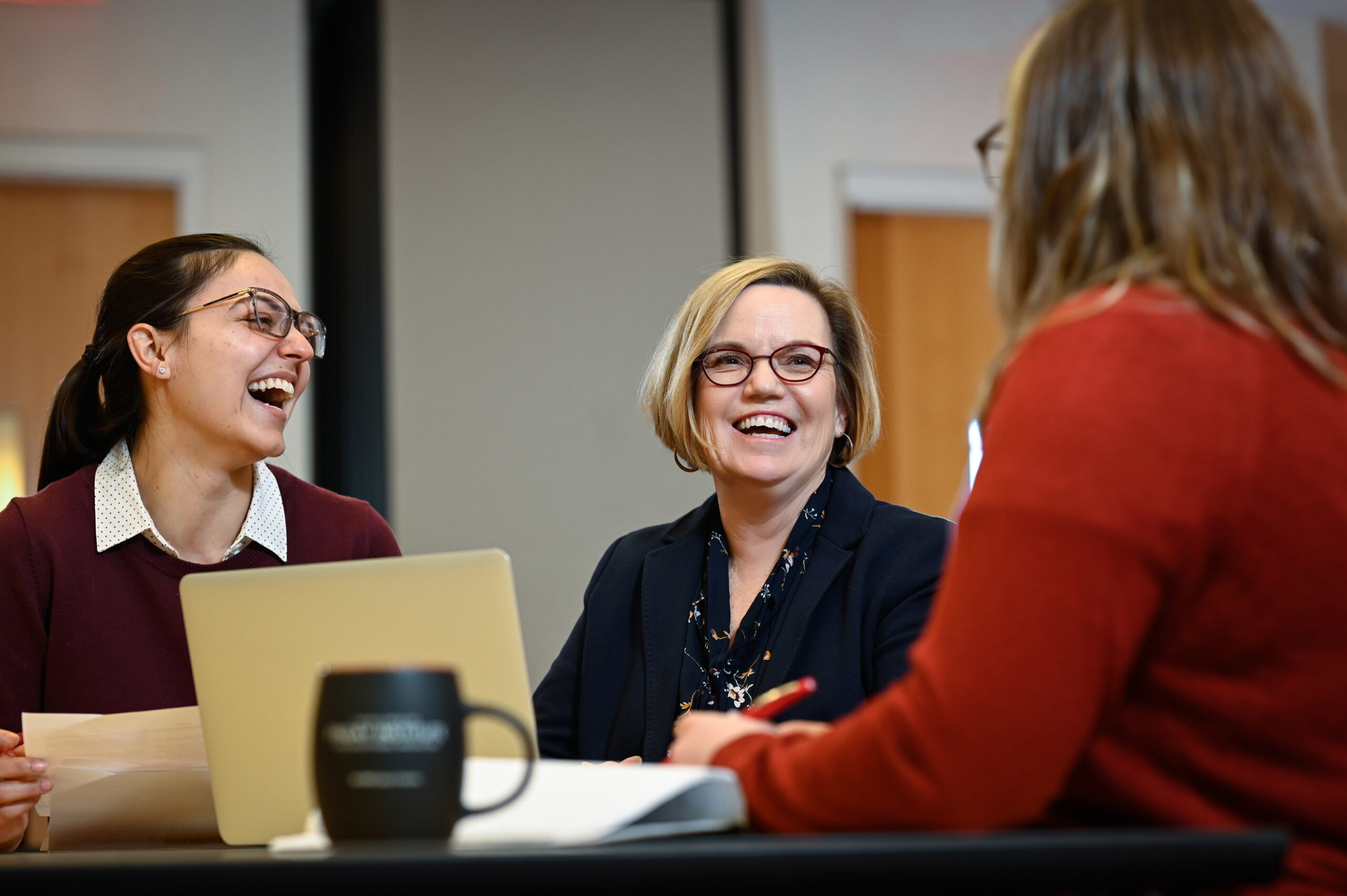 A group of women stand around a table talking and smiling. The photo focuses on a woman with short blonde hair and glasses, Gail Jones.