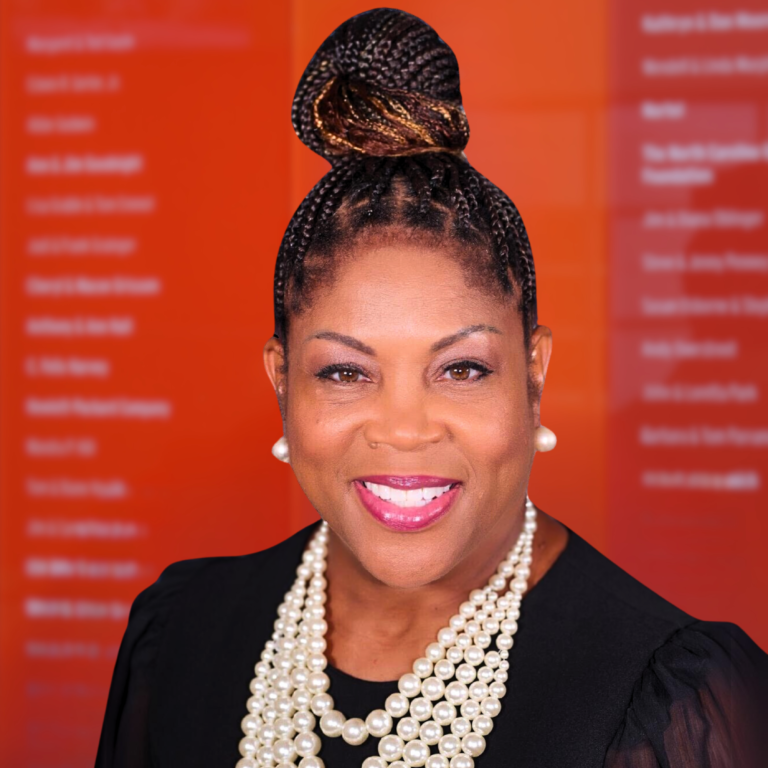 Krista is dressed in black with pearl necklace and earrings. She stands in front of an orange and red background.