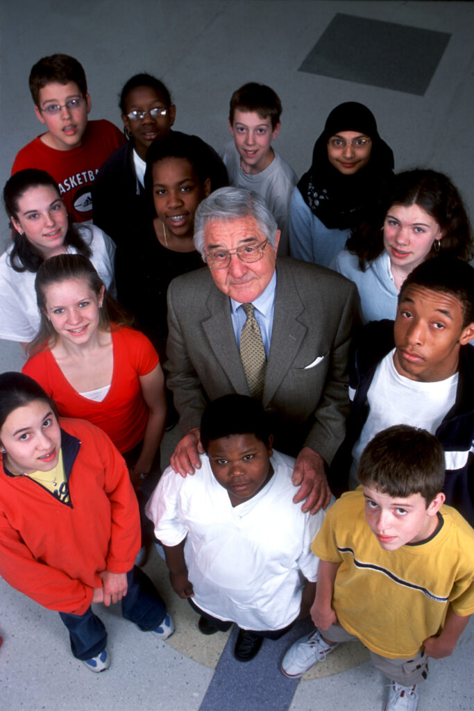 Bill Friay, a man in a suit and tie with gray hair, stands in a cluster of middle school aged students, putting his hands on the shoulders of a boy in front of him