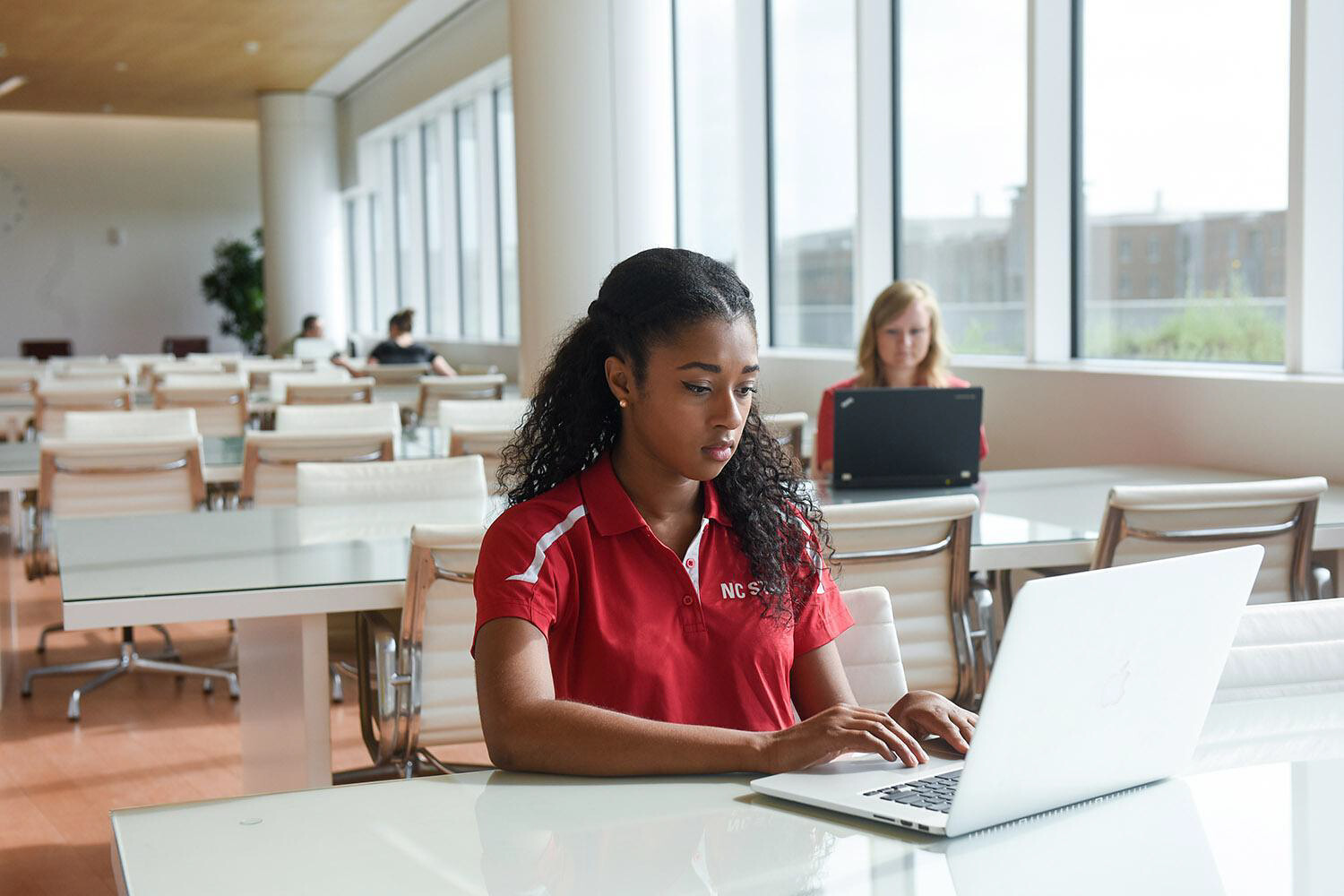 A college student in a red and white and NC State polo shirt sits in front of a laptop at a table in a white room with a wall of windows