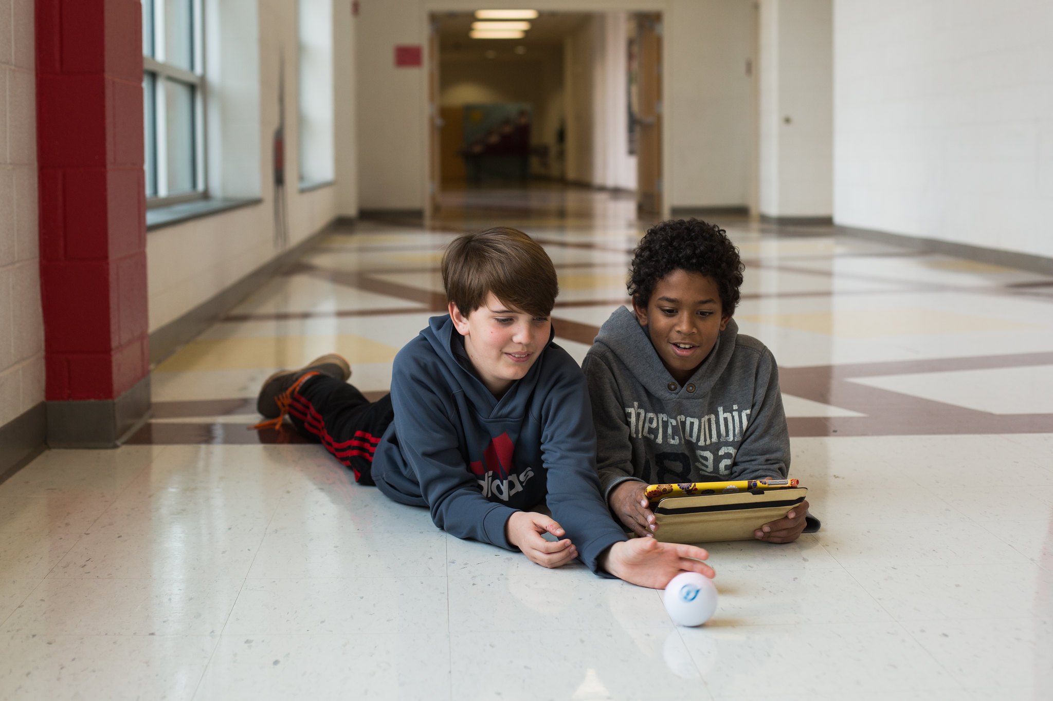 Two students lay on their stomach on the floor of a middle school hallway while playing with a spherical robot.