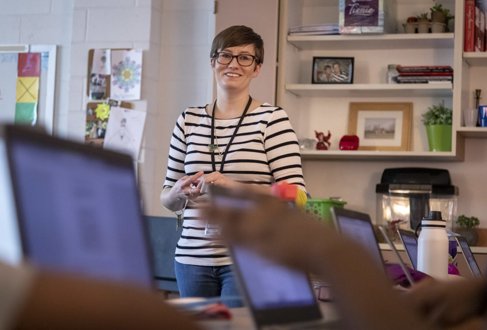 A teacher stands in front of a classroom of students on laptops. She's smiling at the group.