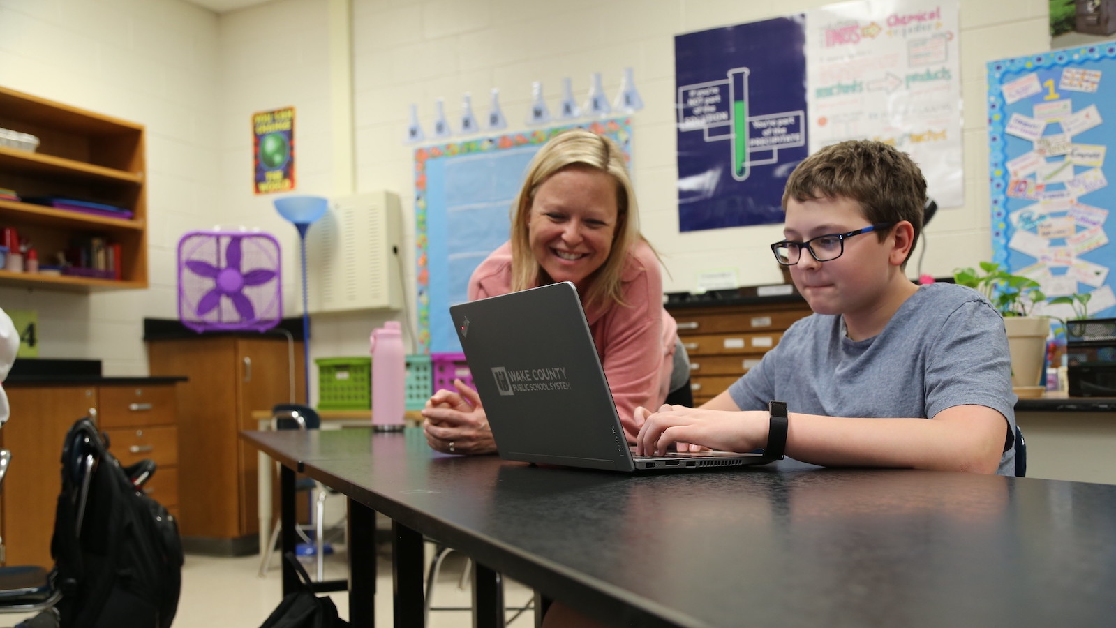 A teacher and student smile as they work on a laptop together in a classroom. The student is sitting at a desk while a teacher leans on the desk.