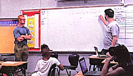 Two Friday Institute male staff stand in front of a white board in a classroom and write on it in front of students.