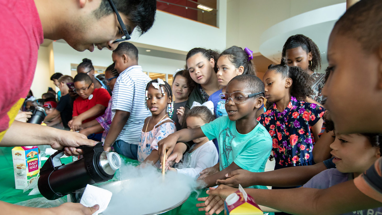 A group of young children stand in front of a silver bowl with steam coming out of it. One child stirs the steam with a spoon while wearing goggles while an adult pours liquid into the bowl.