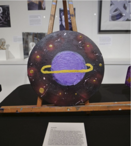 A round painting featuring a planet with a ring around it surrounded by stars.