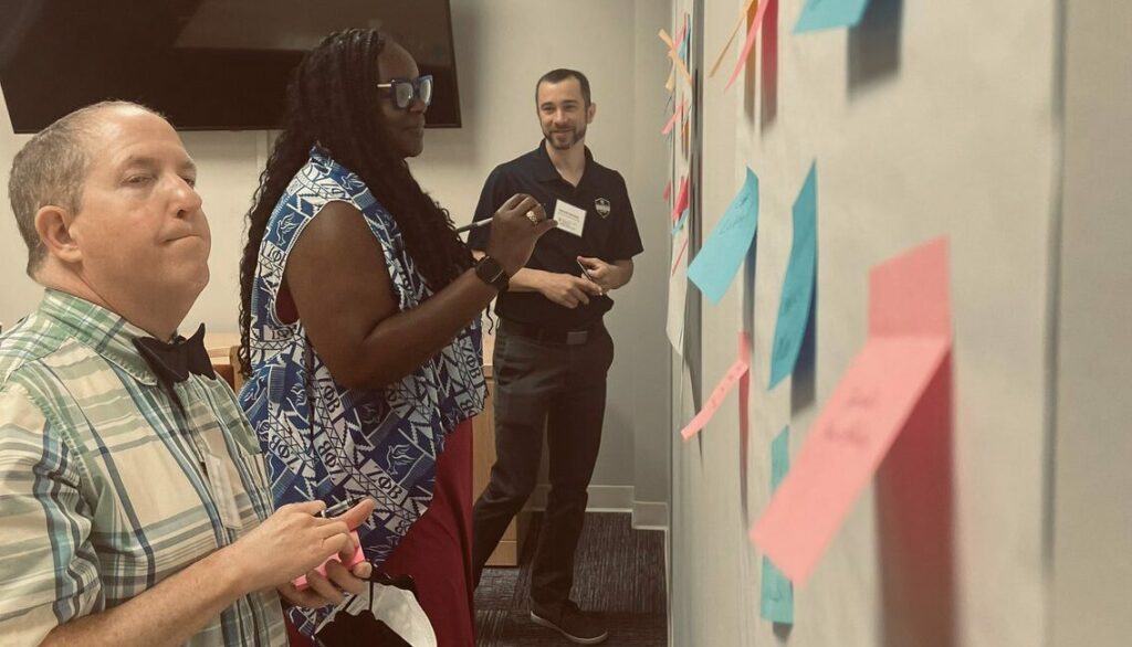 Three educators stand in front of a white board where dozens of different colored post it notes are stuck to the board. The educators are looking at and writing on the post-it notes while talking with each other.