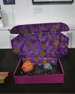 A purple box with stars painted on the top that holds two rocks painted as planets resting on a black sand bottom.