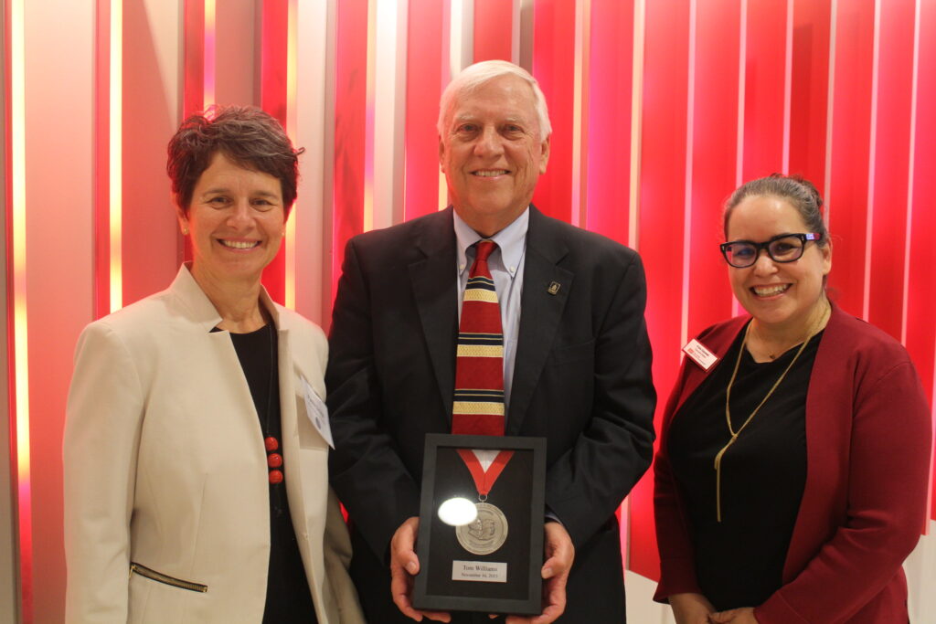 Image of three people: two women on either side of Friday Medal recipient Tom Williams. Tom is a man in a suit and tie holding a medal in a black frame. The women on either side are both wearing blazers and are standing in front of a red background