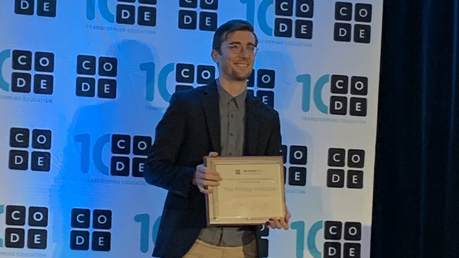 A man with short brown hair and blazer holds up a certificate in from of a Code.org step and repeat background