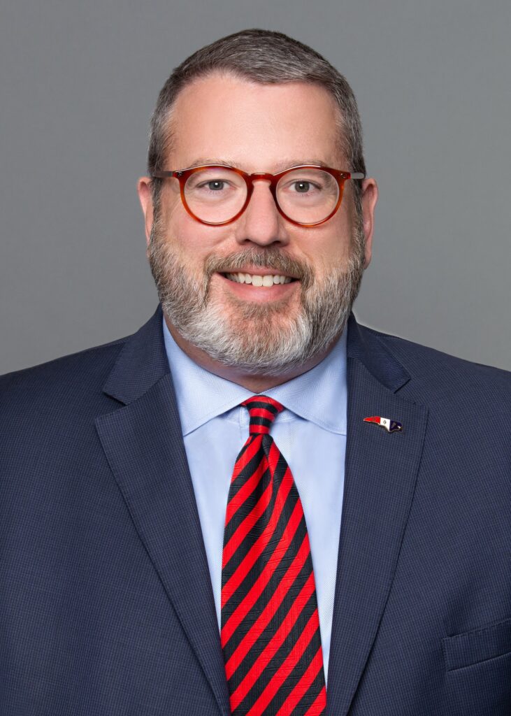 Image of John Denning, a man in a navy suit, blue shirt and red and navy tie. He has a North Carolina state pin on his lapel. He has salt and pepper hair and brown glasses.