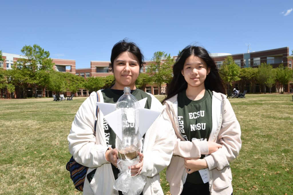 Two students in matching green shirts hold up a science project: a 2-liter bottle with paper fins.