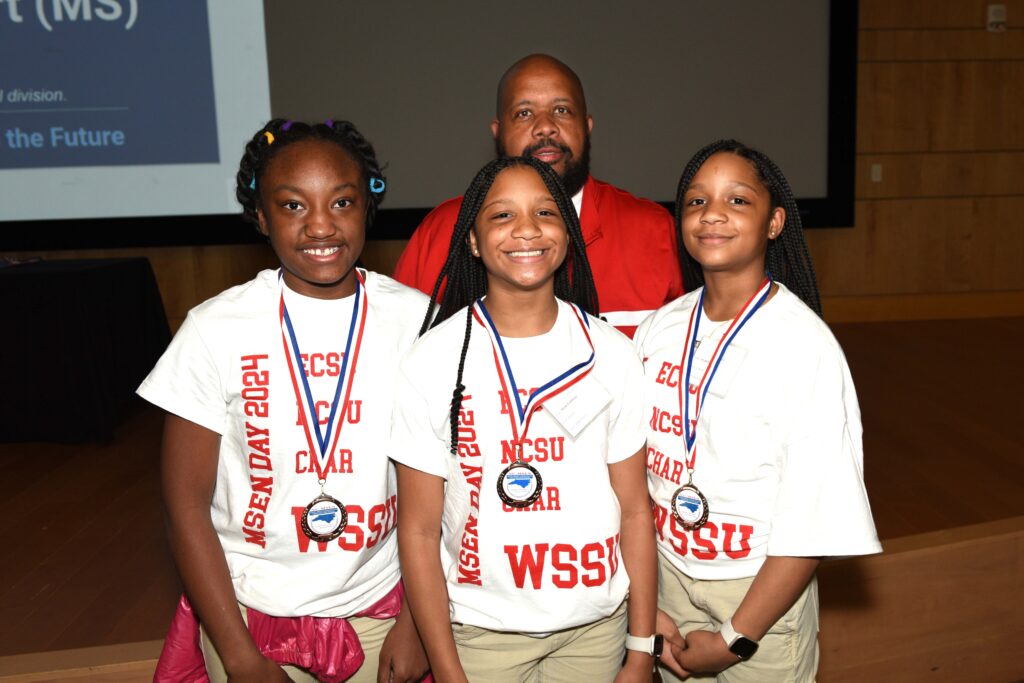Three female students wear white and red t-shirts with their school as the dominant text, WSSU. They re all wearing red, white and blue medals around their necks.