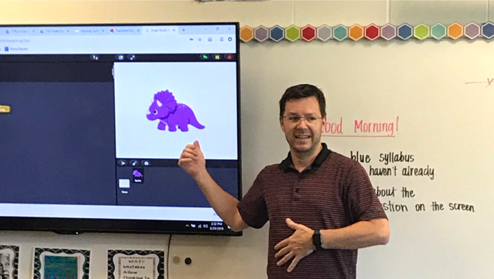 A male teacher stands in front of a dry erase board in a classroom  and pointing at a tv screen with a dinosaur on it as part of a coding app