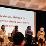 A panel of student women sit in a row on a stage in front of a screen that says "Why do you think it is important to share your story with others?"