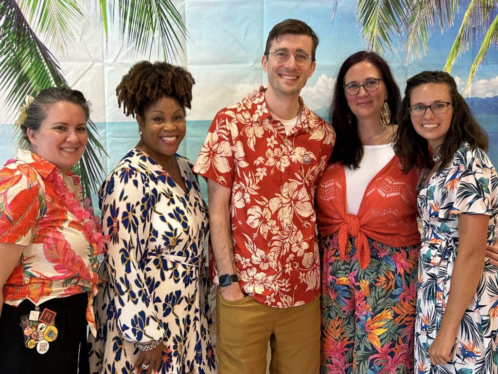 Five people stand in a row wearing Hawaiian print clothing and leis.