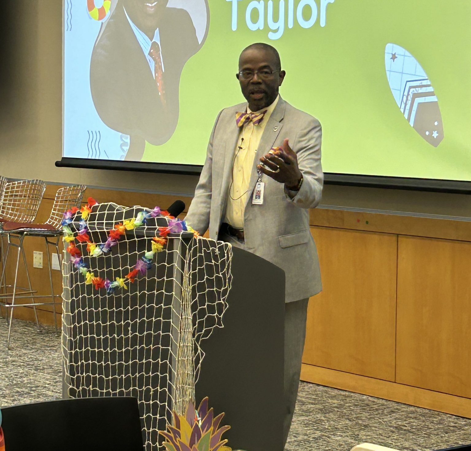 A man stands at a podium in a suit and tie talking to a crowd. The podium is decorated with a lei and fishing net.