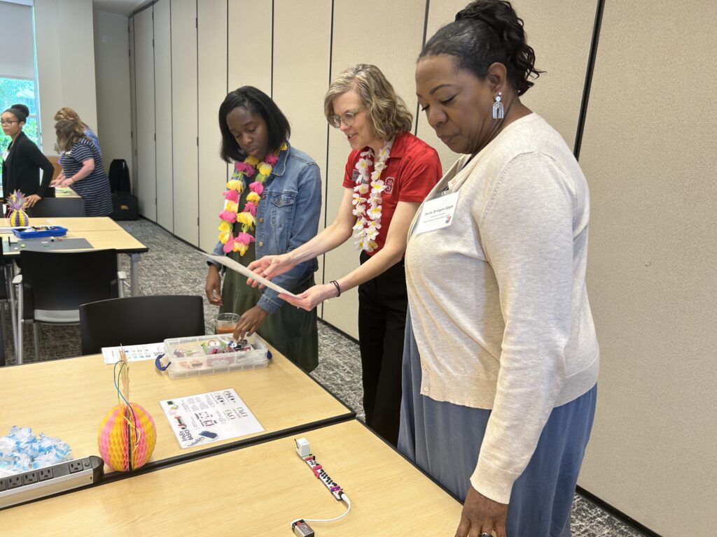 Three women look a a table with a small clear box of circuits.