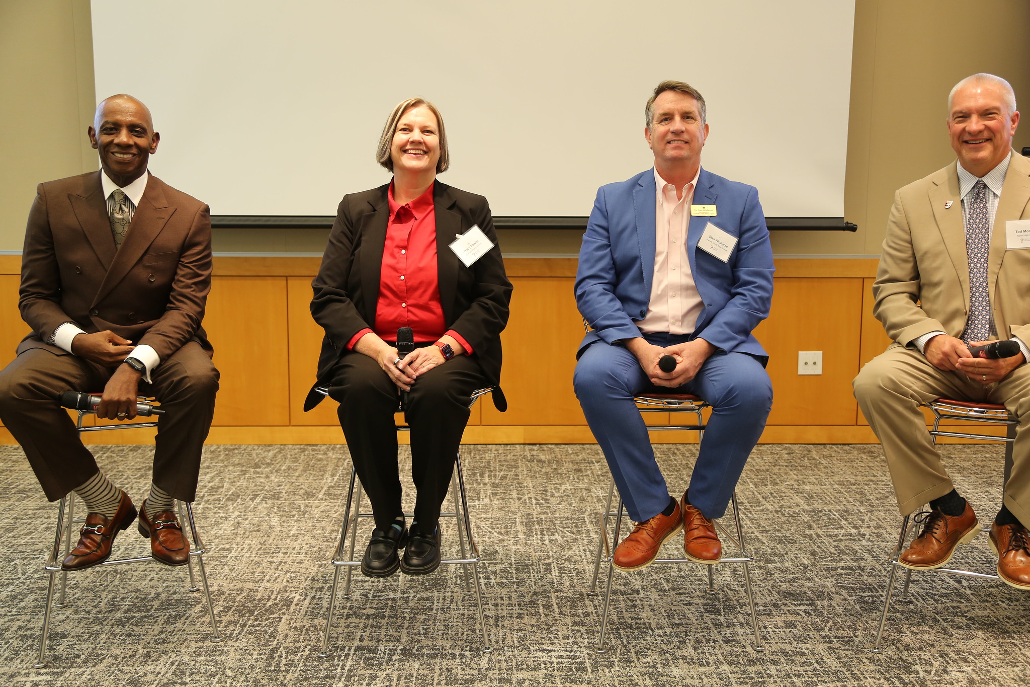 Four educators sit in a row on tall chairs smiling at the camera. They are all wearing suits.