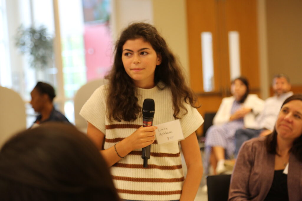 A high school girl in a striped sweater speaks to a room on a microphone.