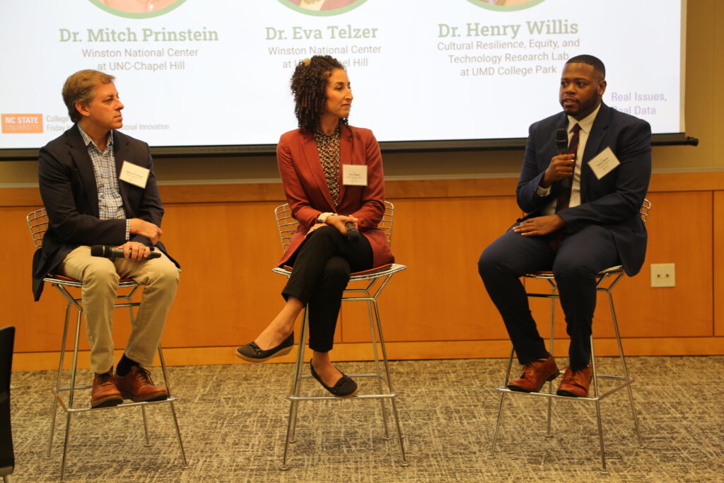 Three people sit on a panel in tall chairs. All wear blazers. One speaks into a microphone while the others listen.