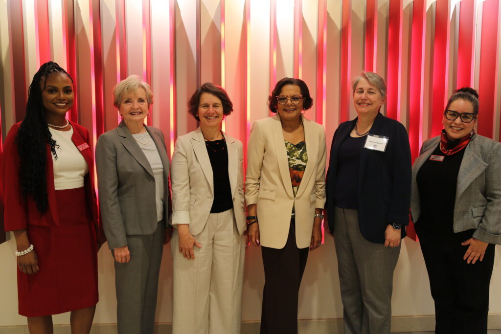 Six women stand in a line in front of a red backdrop. They are all wearing business casual attire and are in blazers.