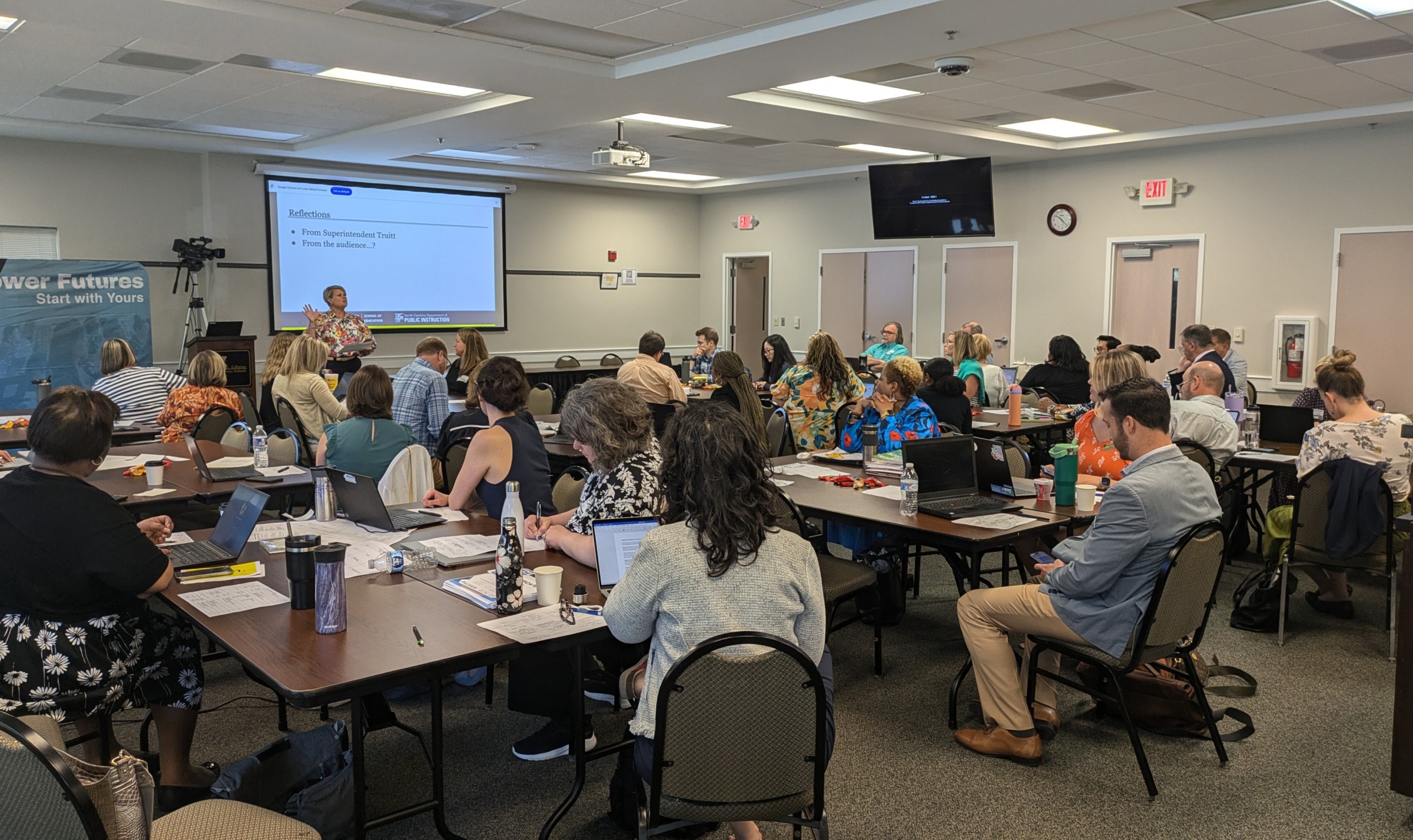 A room of educators sit around tables while a woman presents in the front of the room