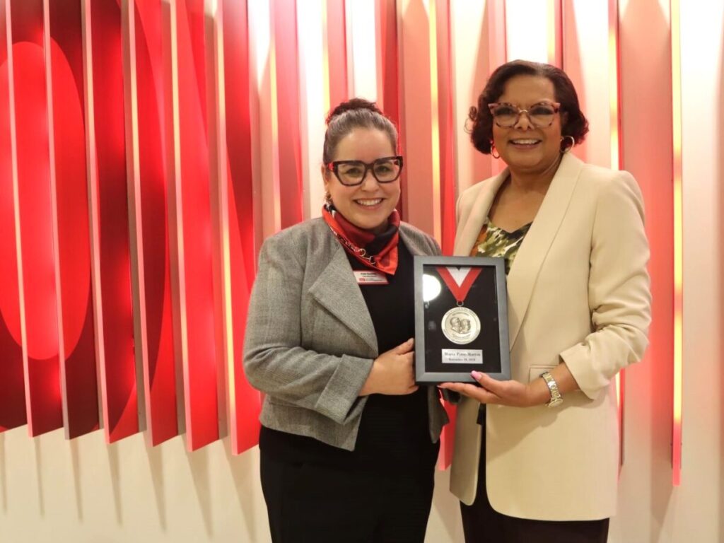 Two women stand in blazers in front of a red backdrop. They hold a frame between them holding a medal hanging on a ribbon.