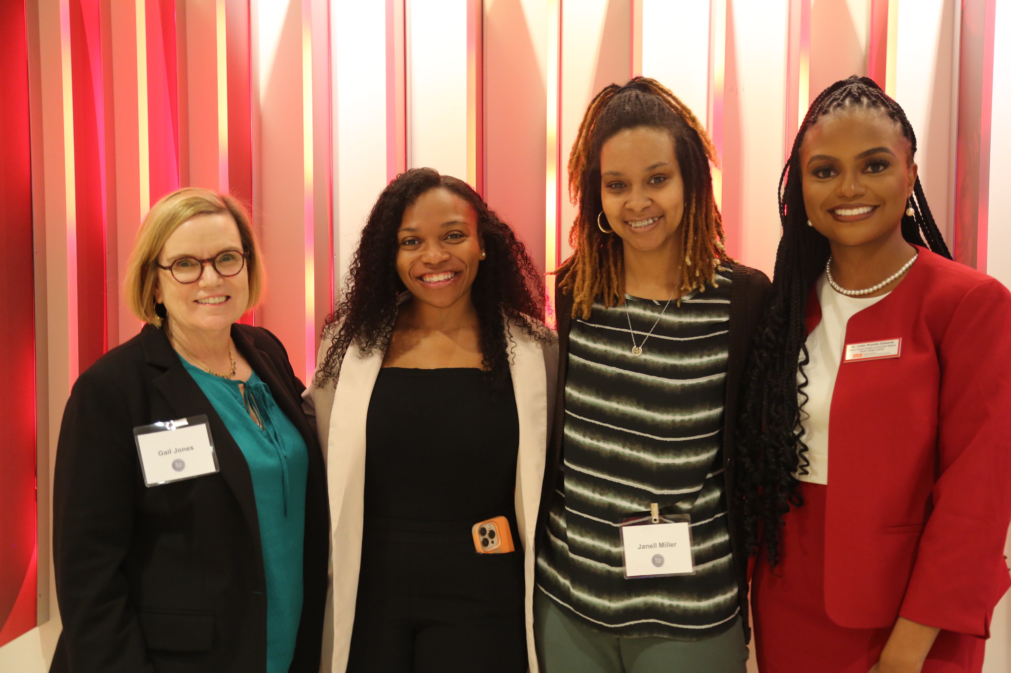 Four women stand in a row in front of a red background.