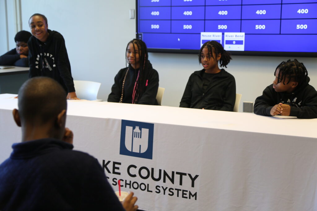 Students sit behind a table with a Wake County Public Schools tablecloth answering questions in front of a jeopardy screen of questions.