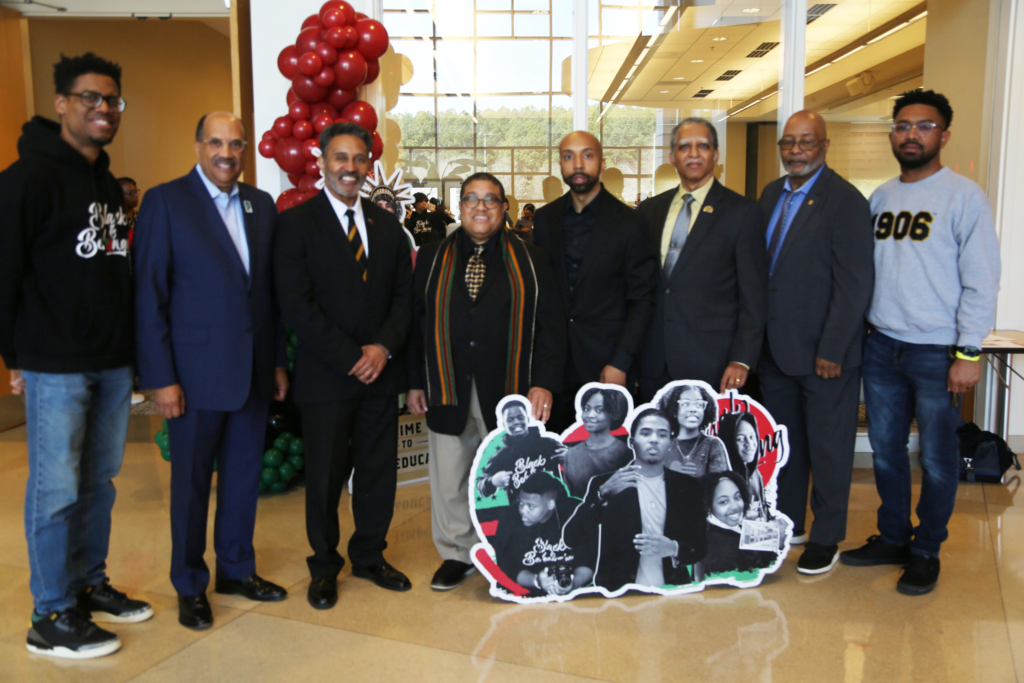 A group of men from the Alpha Phi Alpha Fraternity, Inc. pose together in front of a sign for the event featuring students.