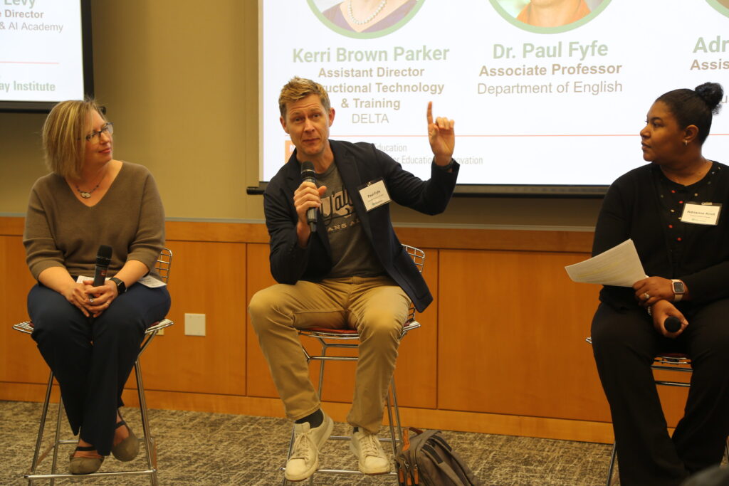 Three panelists sit on high backed stools. The man in the middle holds a microphone while speaking to the audience.