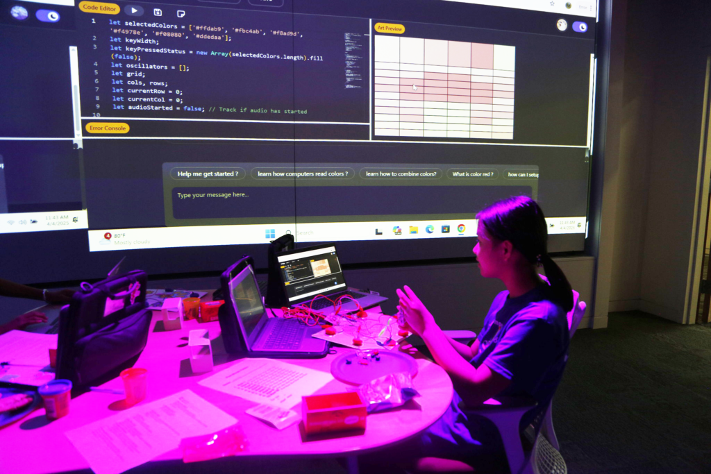 A girl works in front a large screen on the wall displaying computer code. She sits in front of her laptop, which is connected to a bunch of wires in front of her.