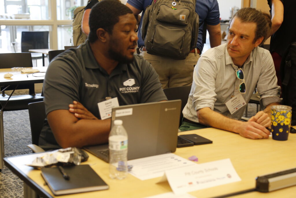 Two professional talk at a table. One sits in front of a laptop.