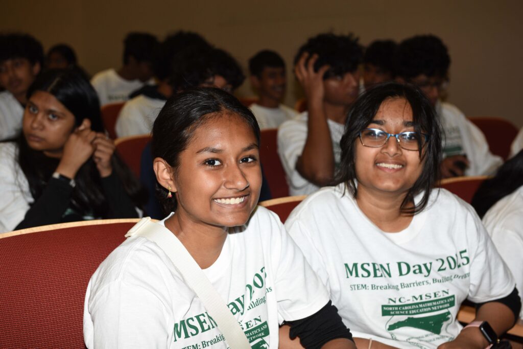 Two students sit in auditorium seats. Several other students are seen in the background.