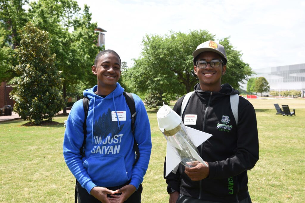 Two students standing outside in a large, grassy open area. One student is holding a model rocket.