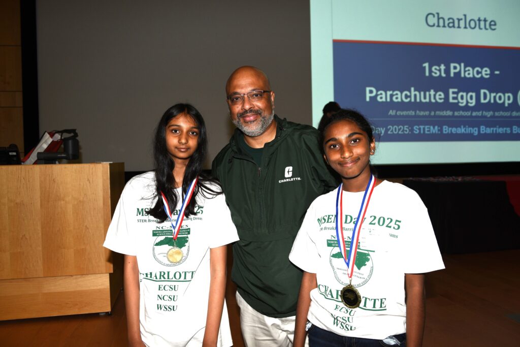A teacher stands in between two students who are wearing medals. They are standing in front of a podium and part of a large screen is visible behind them.