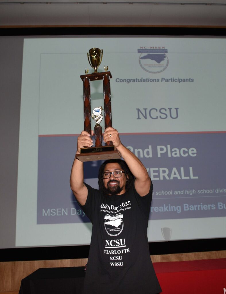 A man stands in front of a large screen holding up a trophy above his head.