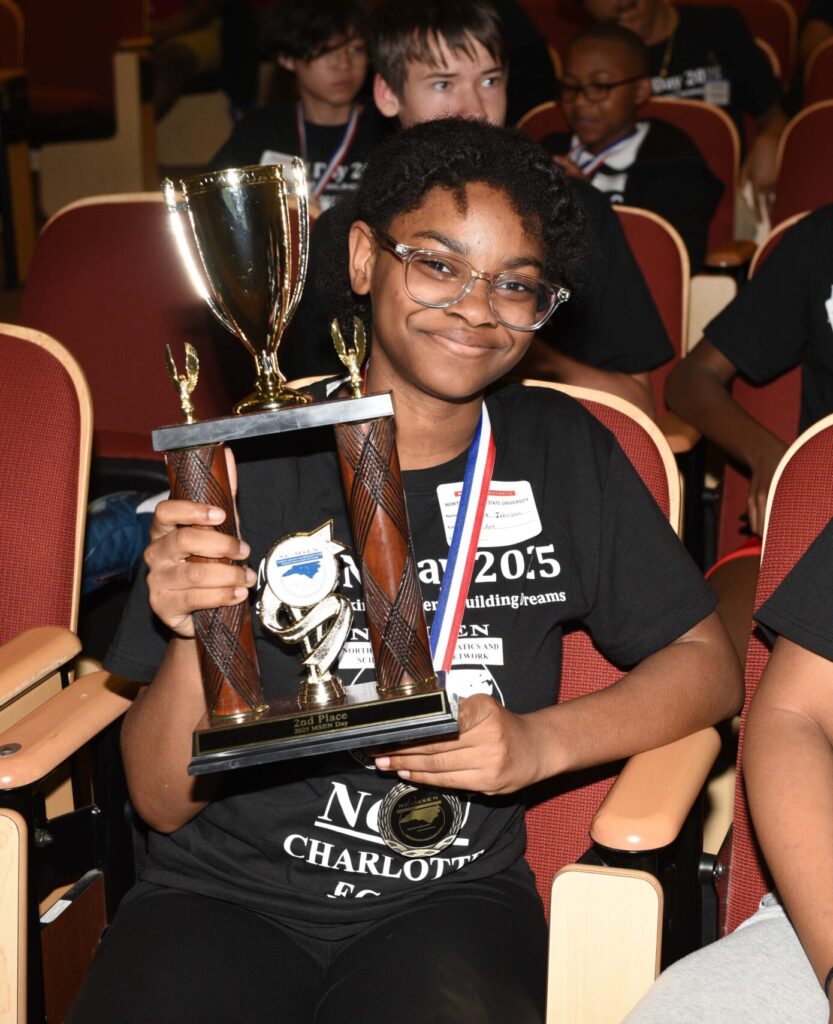 A student sits in an auditorium with other students in the background. The student holds a trophy.