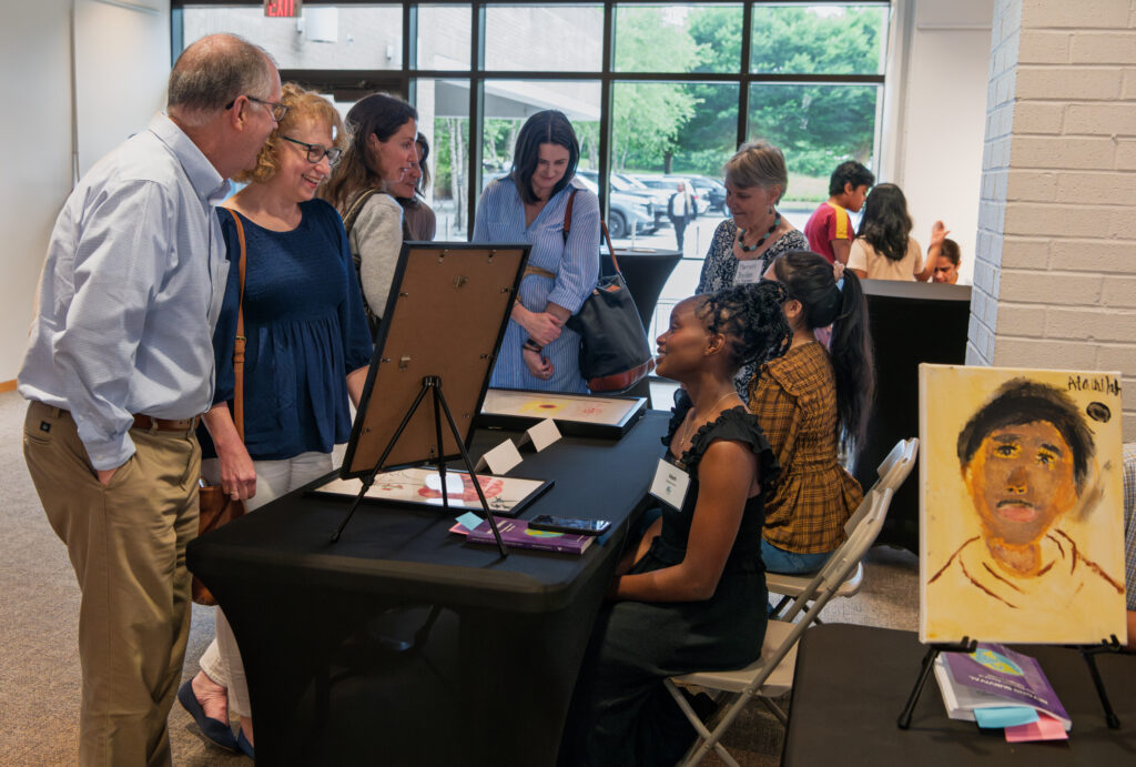 A girl sits at a table in a crowded room speaking to people standing in front of her table. The table has an easel on it.