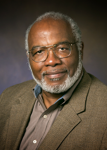 A man with white hair and a beard and glasses wearing a tan blazer and collared button up shirt