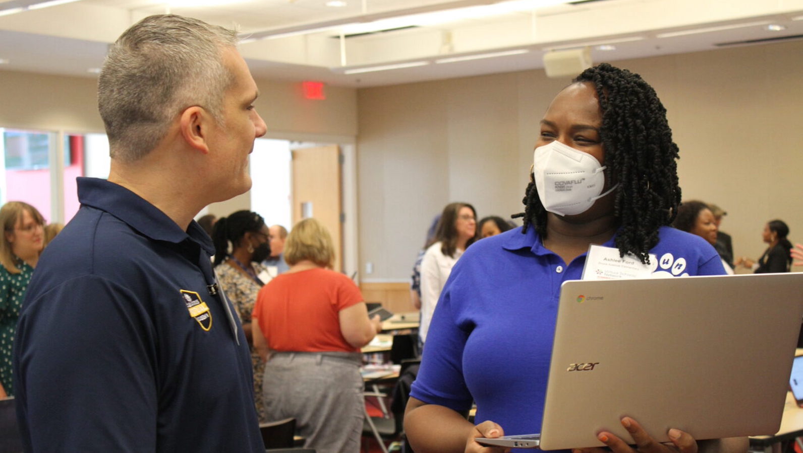 Two teachers converse in a room. One wears a mask and holds a laptop. The other is smiling. Both wear blue polo shirts.