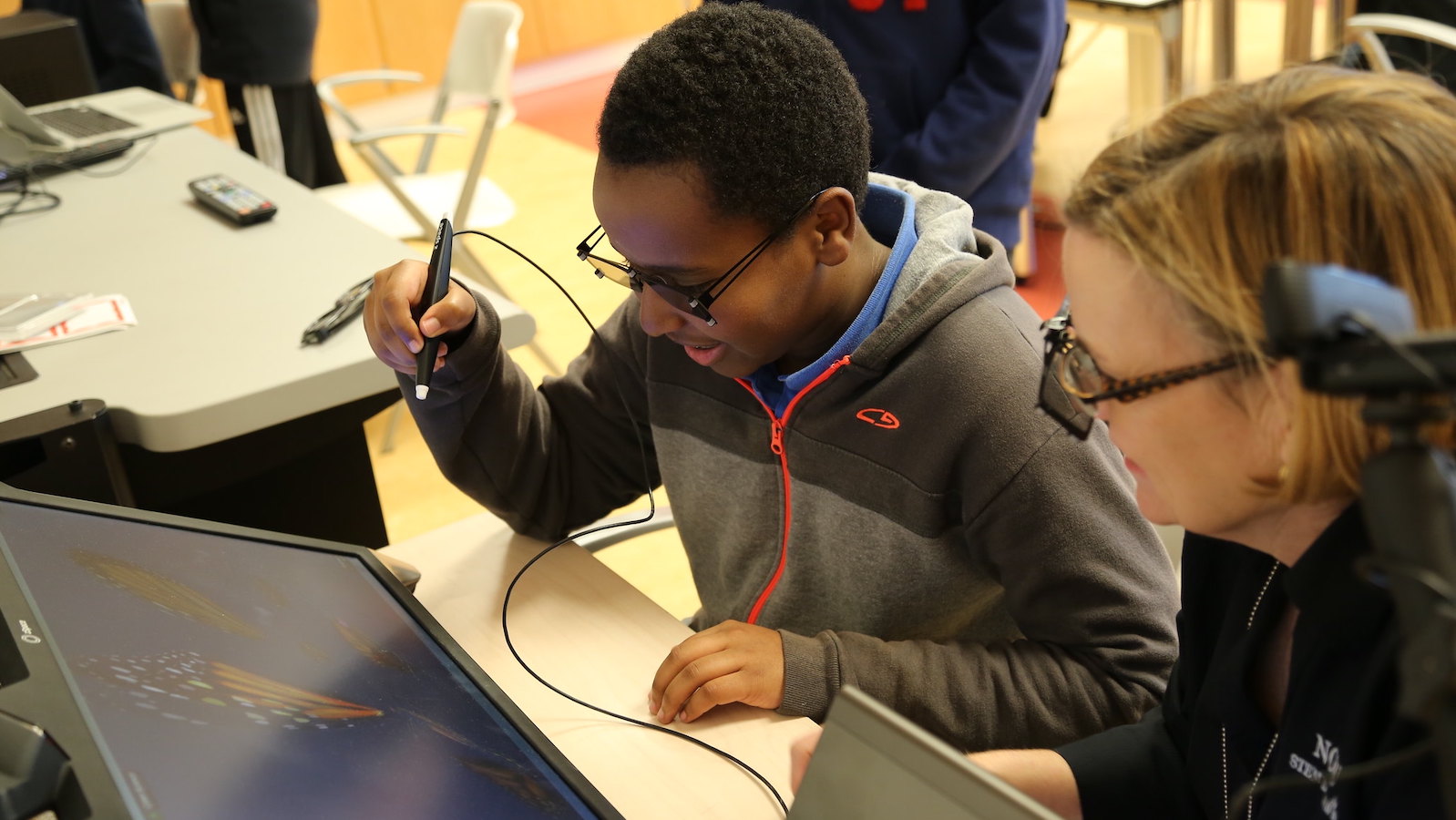 A teacher and student sit at a table over a screen. The student holds a stylus in his hand.