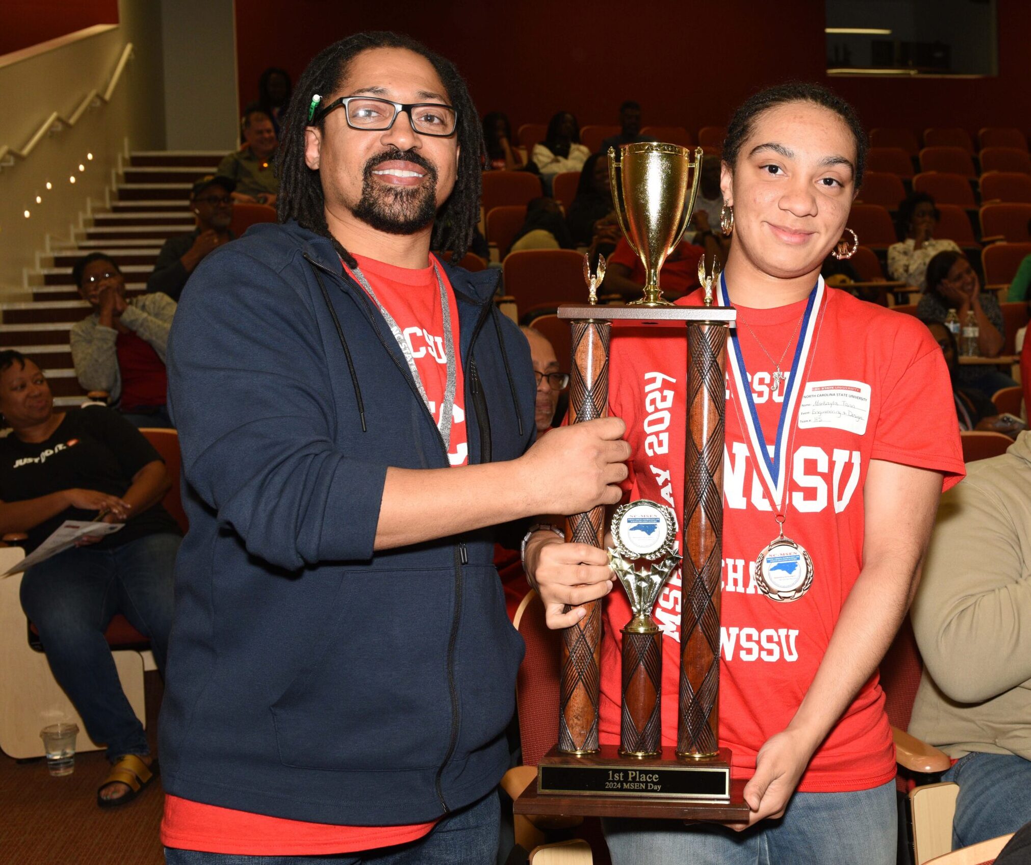 Two people hold up a large red and gold trophy. One is a student. One is an adult. They are both wearing red shirts, as part of the same team.