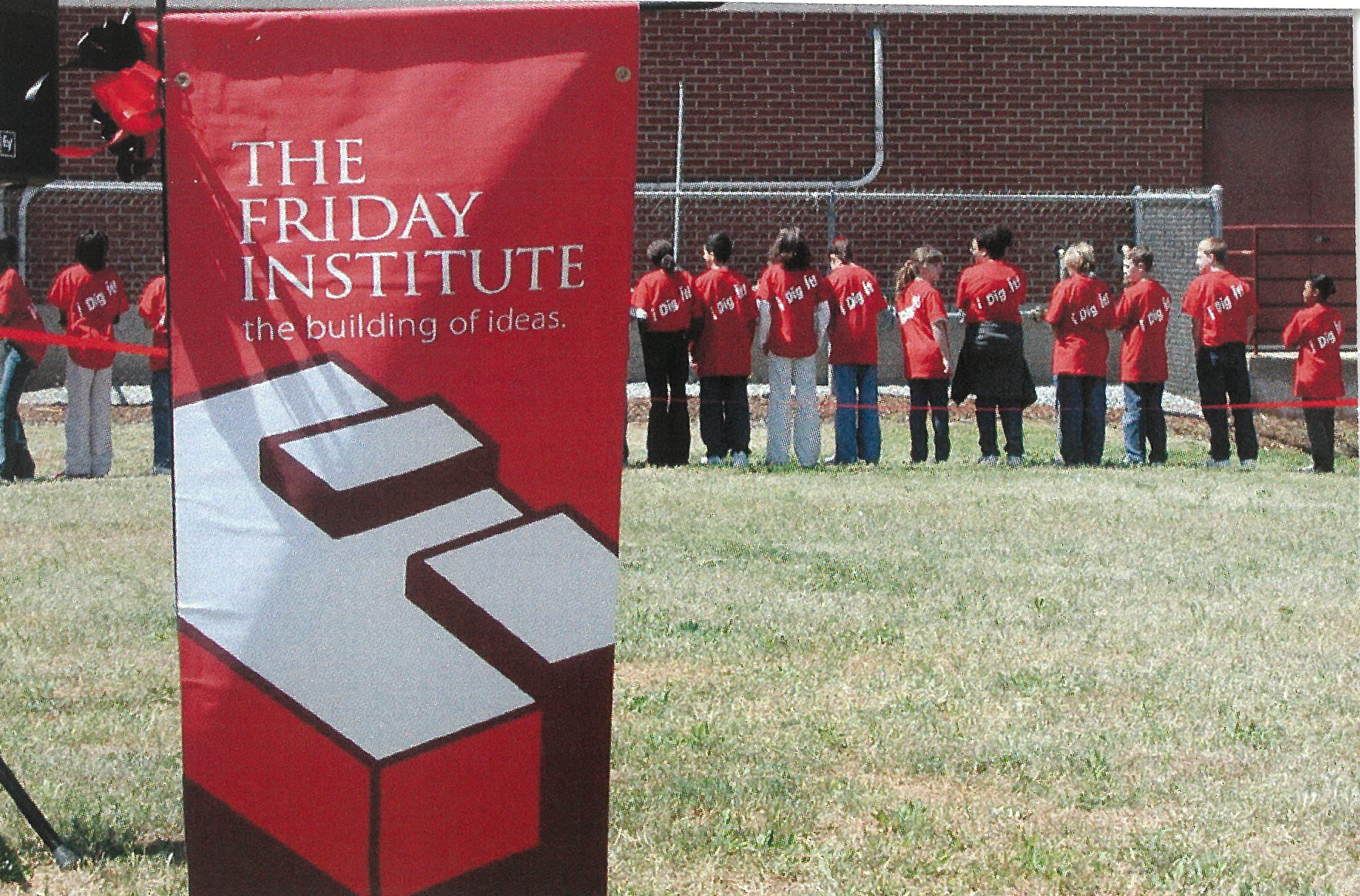 A line of students in red shirt stand with their backs toward the camera in the background. In the foreground, a Friday Institute banner that calls it the Building of Ideas