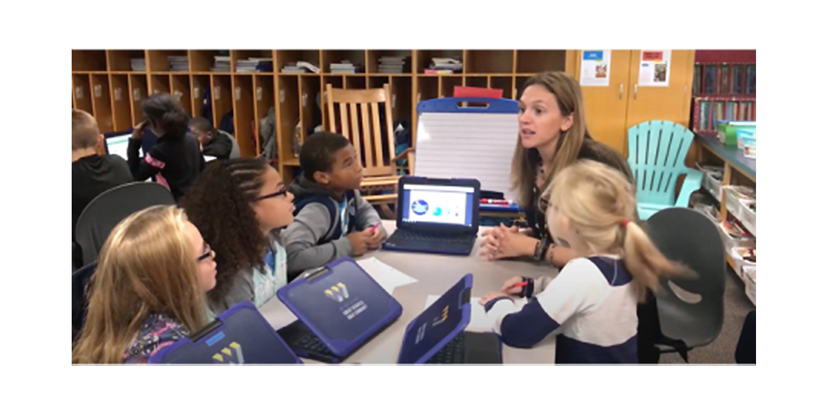A teacher in a classroom speaks to students at a table while they listen in front of tablets and paper